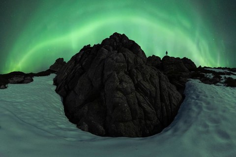 Unrecognizable person silhouette standing atop rugged snowy rocks under a vibrant aurora borealis