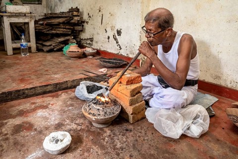 Man uses a blowpipe to spark a coal-fired vessel, a man burns sapphires in a small stove in the