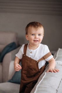 A joyful toddler smiles brightly while standing in a cozy, softly lit living room. Wearing brown