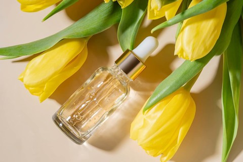 A glass dropper cosmetic of luxurious skincare serum rests among vivid yellow tulips on a soft