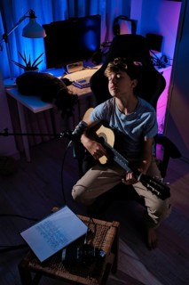 A young musician sits with a guitar in a cozy lit room. A notebook and recording equipment are