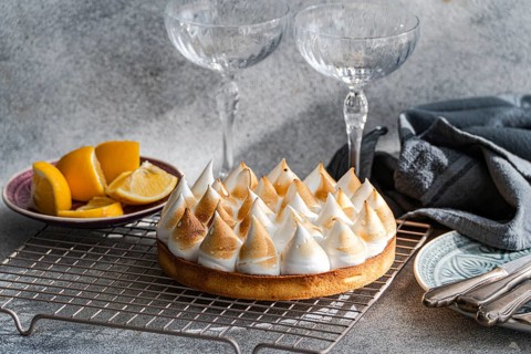 A homemade lemon cake on a cooling rack beside sliced lemons. Two empty glasses add a touch of