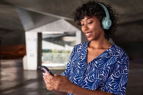 A woman wearing blue headphones smiles while looking at her phone, standing in a modern