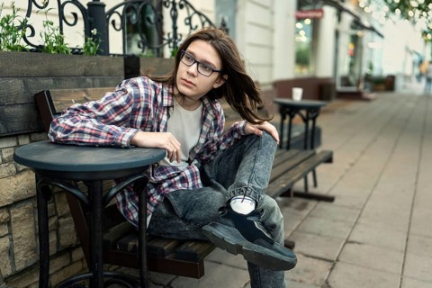 Teenage boy with long hair and glasses sitting on a bench in an urban setting, wearing a checked