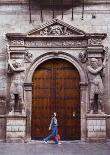 A young woman walks by an ornately carved wooden entrance, framed by sculpted stone figures. The