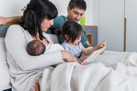 A young family sits together on a cozy bed. The parents watch with smiles as their daughter, a big
