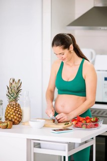 A pregnant woman in a green yoga outfit stands in a kitchen slicing strawberries. A variety of