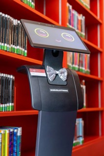 The robot stands in a library next to red bookshelves, interactive guided tours by robots