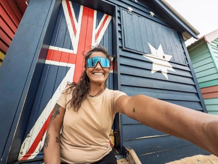 A cheerful woman takes a selfie in front of the iconic Brighton Bathing Boxes in Melbourne,