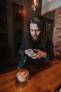 A bearded man intently uses his smartphone while sitting at a wooden counter in a cozy cafe. A cup