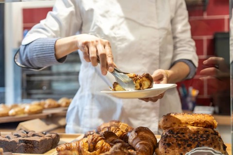 Baker wearing an apron and blue sweater serving a piece of chocolate pastry from a display case