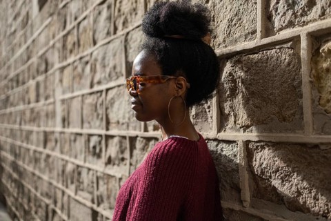 A stylish woman in a burgundy sweater wearing sunglasses stands confidently against a textured