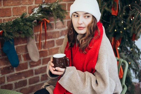 A young woman in a white beanie and red scarf holds a hot drink, seated outdoor on a snowy day,