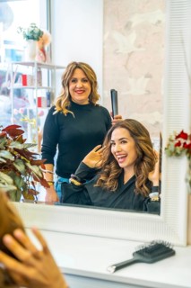 Hairdresser styling woman client's long curly hair with a flat iron while both women smile,