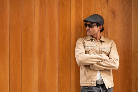 A Colombian gay man wearing sunglasses and a flat cap stands confidently against a wooden wall with