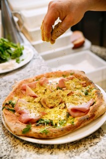 Cropped unrecognizable person sprinkling seasoning on a homemade pizza topped with ham and arugula,