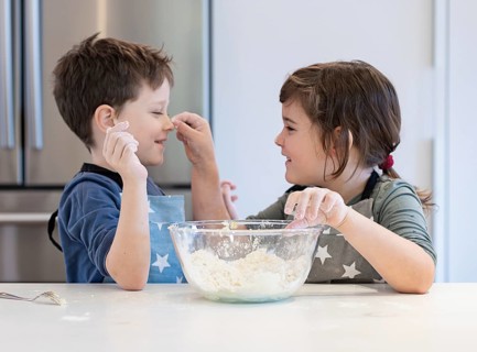 Two children wearing aprons enjoy making dough in a kitchen. They laugh and playfully interact,