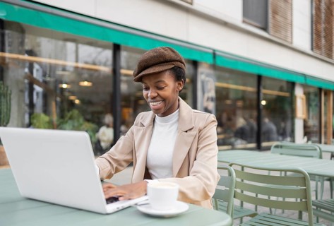 A black woman smiles while working on her laptop at an outdoor cafe Her stylish outfit and relaxed