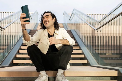 A young man with curly hair and a mustache wearing casual attire sits on outdoor steps, holding a
