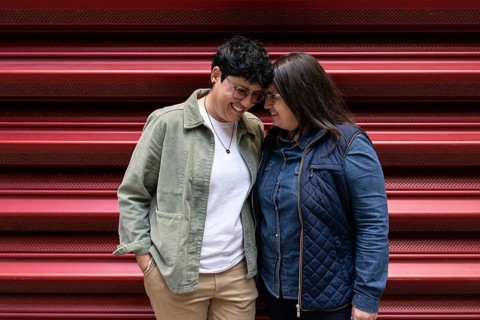 A curvy lesbian couple share a light heart moment, smiling warmly as they lean into each other.
