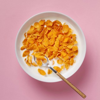 Top view of a white bowl filled with golden cornflakes and milk, accompanied by a metal spoon, set