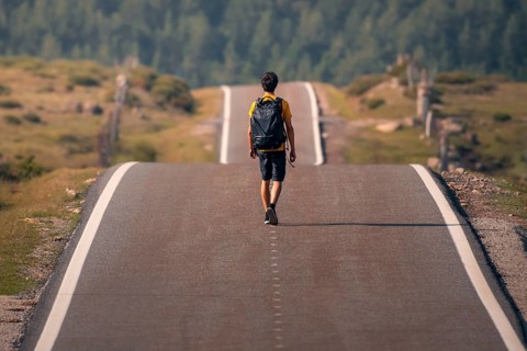 A solitary man with a backpack walks down a deserted winding road through verdant scenery in