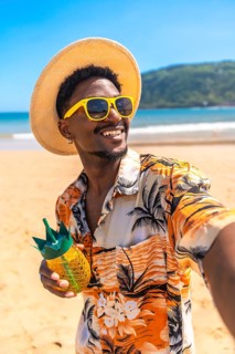 Young man wearing sunglasses and straw hat enjoys a tropical drink on a sunny beach while taking a