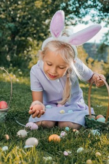 Young girl with bunny ears collects colorful Easter eggs in a sunny garden. She kneels on the