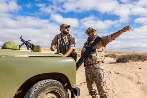 Two soldiers in desert gear strategies near an armored vehicle, one pointing ahead while the other