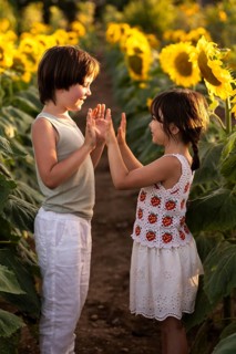 Two children play joyfully in a vibrant sunflower field during sunset. They touch hands, smiling