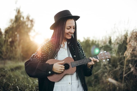 A woman with braided hair and a hat strums a ukulele while standing in a tranquil outdoor setting