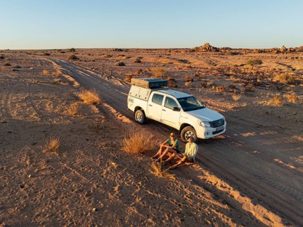 Two travelers sit next to an off-road car on a dirt road in the desert, Erongo, Damaraland, Namibia