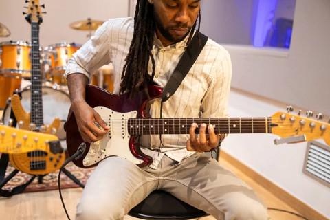 A black musician plays an electric guitar in a professional recording studio, with drums in the