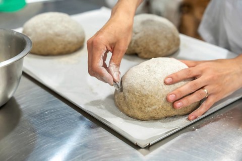 Baker's hands scoring round sourdough loaf with a sharp lame on parchment, close up showing artisan