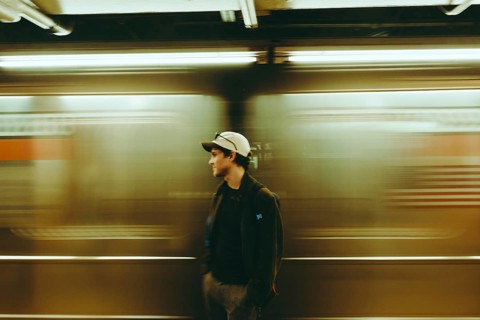 A man wearing a cap stands still on a subway platform while a train speeds by, creating a dynamic