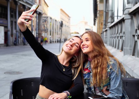 Two teenage girls happily taking a selfie together. They are seated outdoors in a city setting,