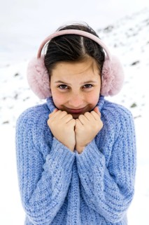 A young girl in a blue sweater and pink earmuffs stands in a snowy winter landscape. She smiles
