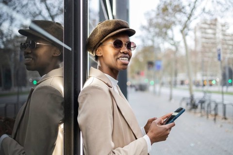 A stylish black woman in a hat and sunglasses smiles while using her smartphone She leans against a