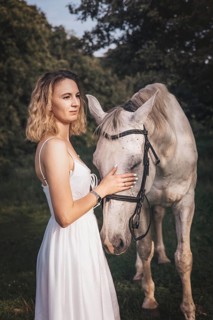 A young woman in a flowing white dress stands in a grassy area, gently caressing a white horse. The