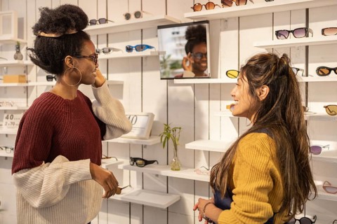 Two women engage happily in a sunglasses store. The customer tries on a stylish pair, enjoying a