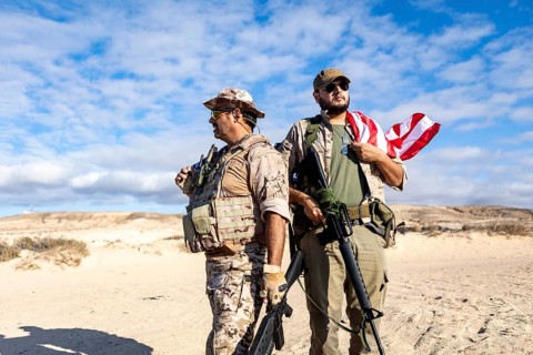 Two armed soldiers stand confidently in a desert landscape. One holds a rifle and dons military