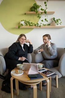 Two women sit comfortably in a stylish cafe, engaged in conversation while using smartphones,