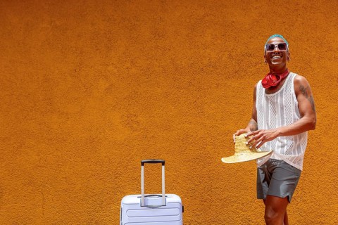 A cheerful LGBTQIA+ man stands with a suitcase against a warm, sunny backdrop. Ready for a holiday,