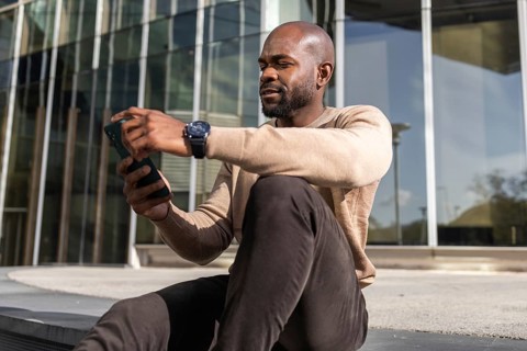 A cuban man sitting outside a modern building, intently looking at his smartphone. He wears a