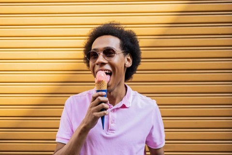 A smiling non-binary person in sunglasses enjoys a delicious ice cream cone against a bright