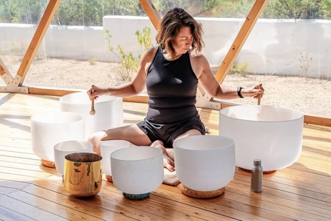 A woman plays crystal singing bowls in a serene wooden structure, using mallets to create soothing