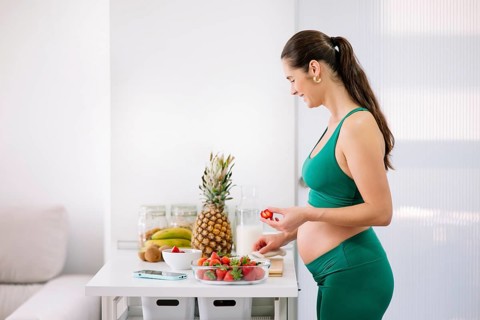 A pregnant woman in a green yoga outfit prepares a fresh, healthy meal in a bright kitchen,