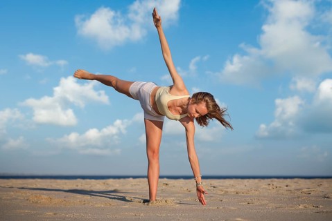 A woman balances on one leg performing Ardha Chandrasana on a sandy beach during sunset with a