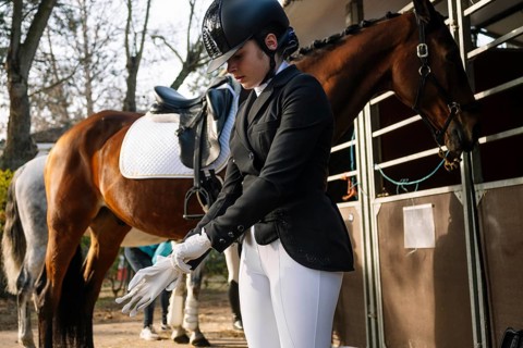 A teenage girl in traditional dressage attire prepares her gloves, with a keen focus outside the