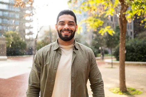 A bearded man smiles warmly under light rain in a park of the city. Sunlight filters through trees,
